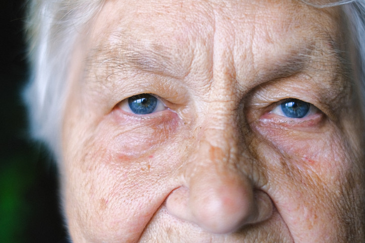 Äldre persons ansikte i närbild. Personen tittar rakt in i kameran med blå ögon och neutral blick.