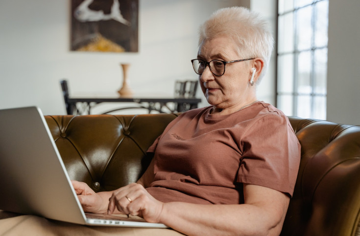 Äldre kvinna sitter i en brun skinnsoffa med en laptop i knät.