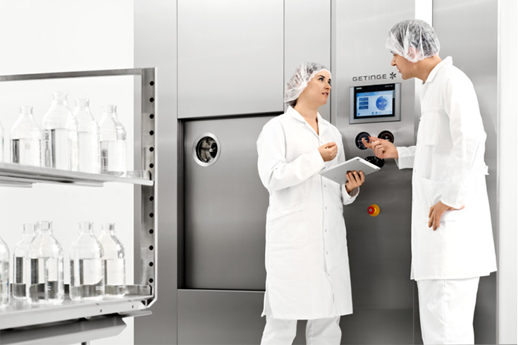 Två personer i laboriatorieklädsel står framför en maskin i blankt stål och pratar. 