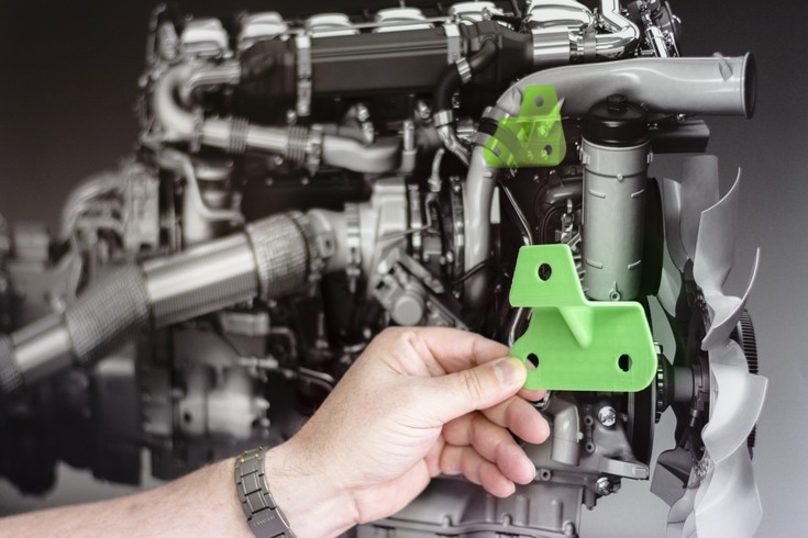 En hand som håller en grön plastform mot en bild av en motor där motsvarande form finns marjerad.