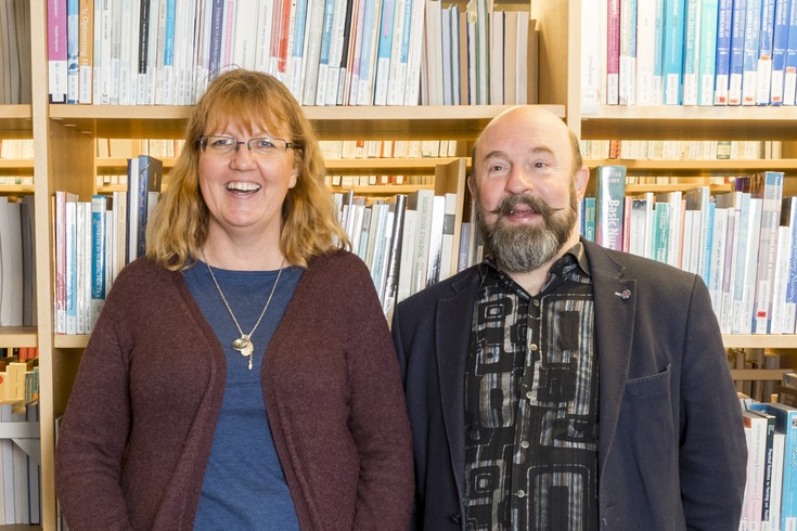 Anna Fåhraus, universitetslektor i engelska, och Jörgen Öijervall, universitetsadjunkt i omvårdnad, skrattar mot kameran, framför hyllor med böcker