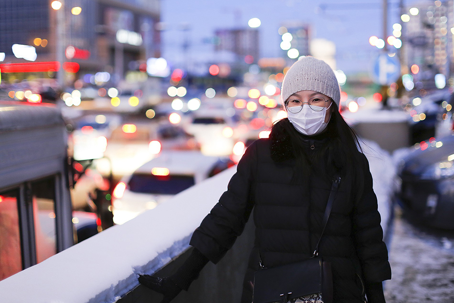 Kvinna med ansiktsmask står vid en mur. En stad och trafik syns i bakgrunden.