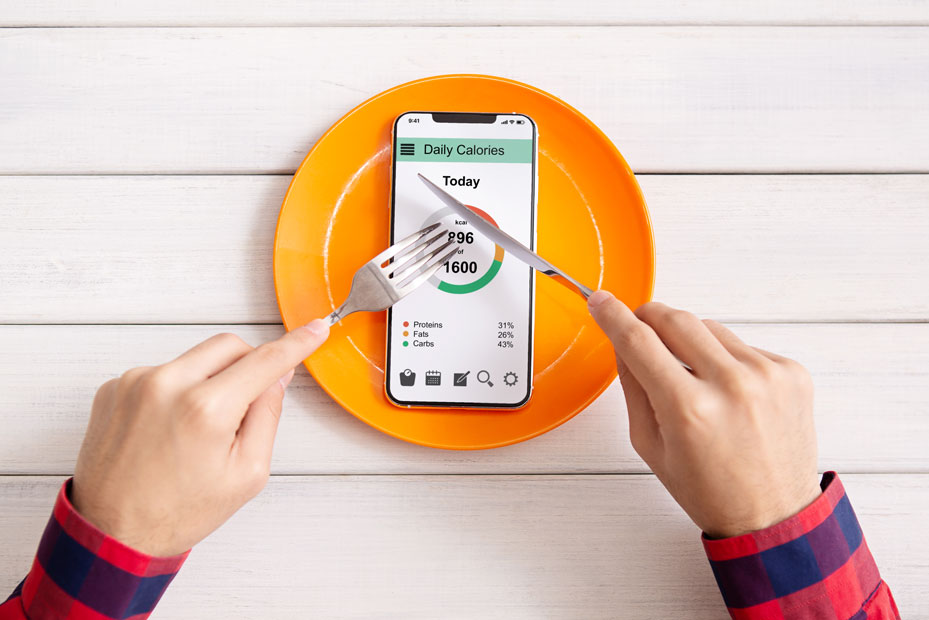 Händer, bestick – redo att hugga sin i en smart telefon, som visar en kalori-app, liggande på en orange tallrik.